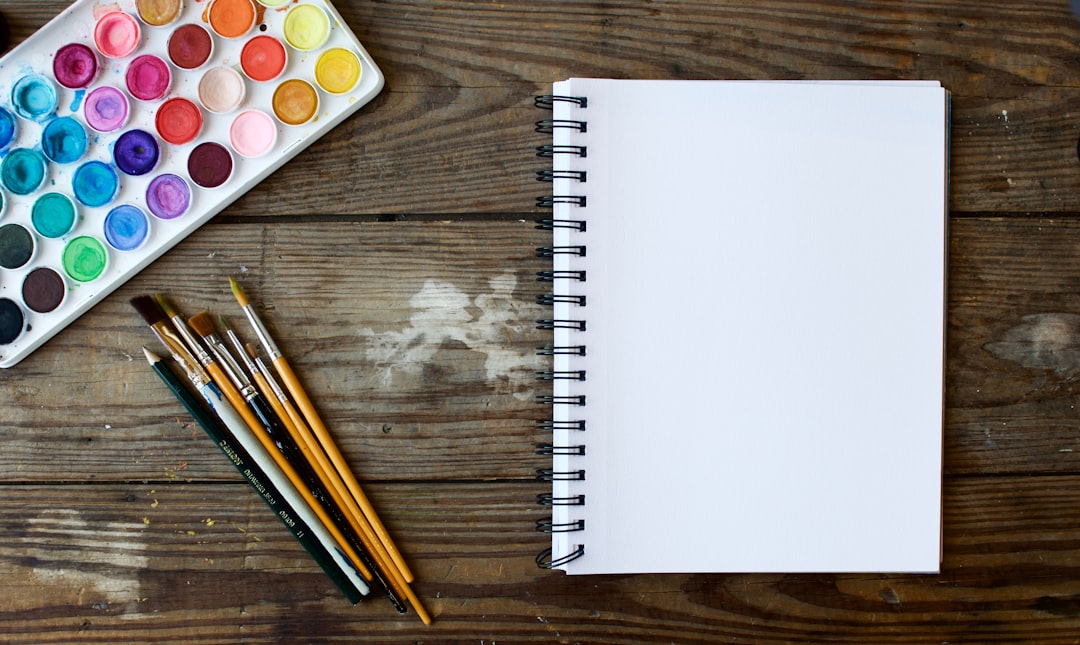 Watercolor palette, brushes, and a blank spiral sketchbook on a wooden table.