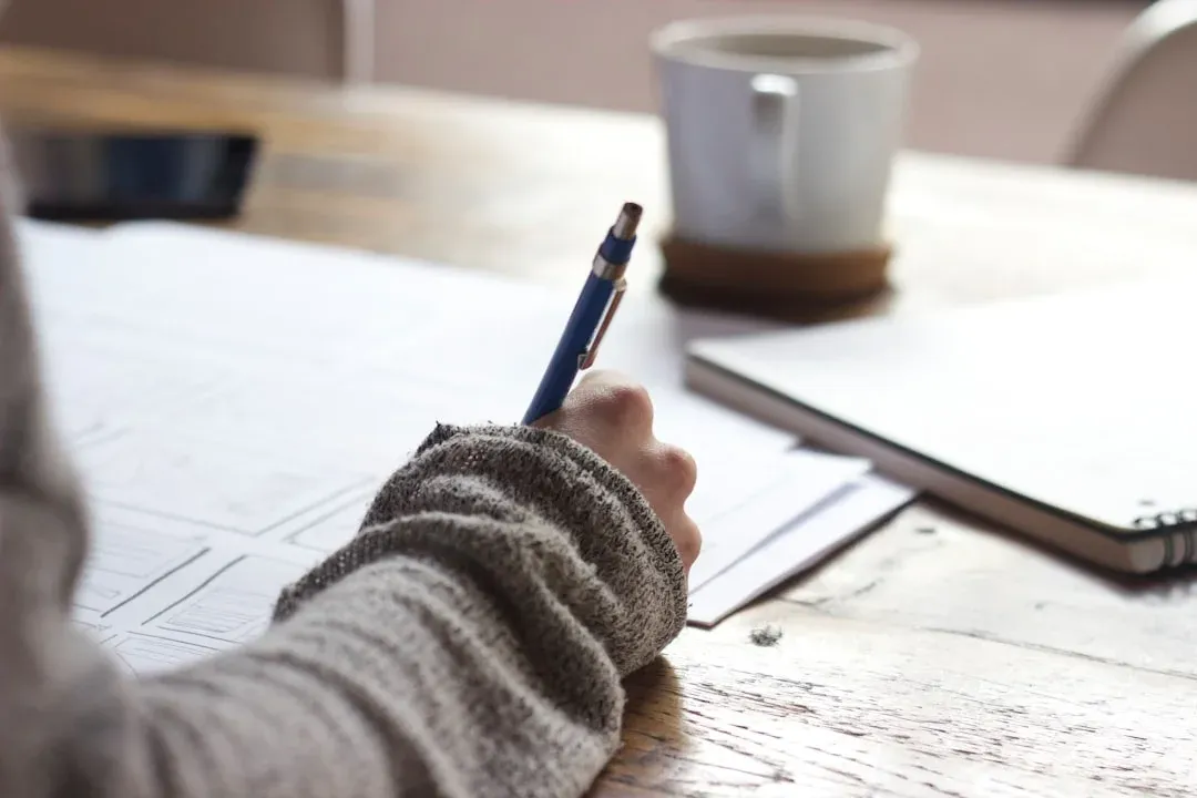 A person writing on papers at a wooden table, with a notebook and a cup of coffee in the background.