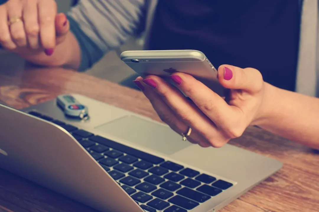 Person with pink nails using a smartphone, with a laptop and USB drive on a wooden desk.