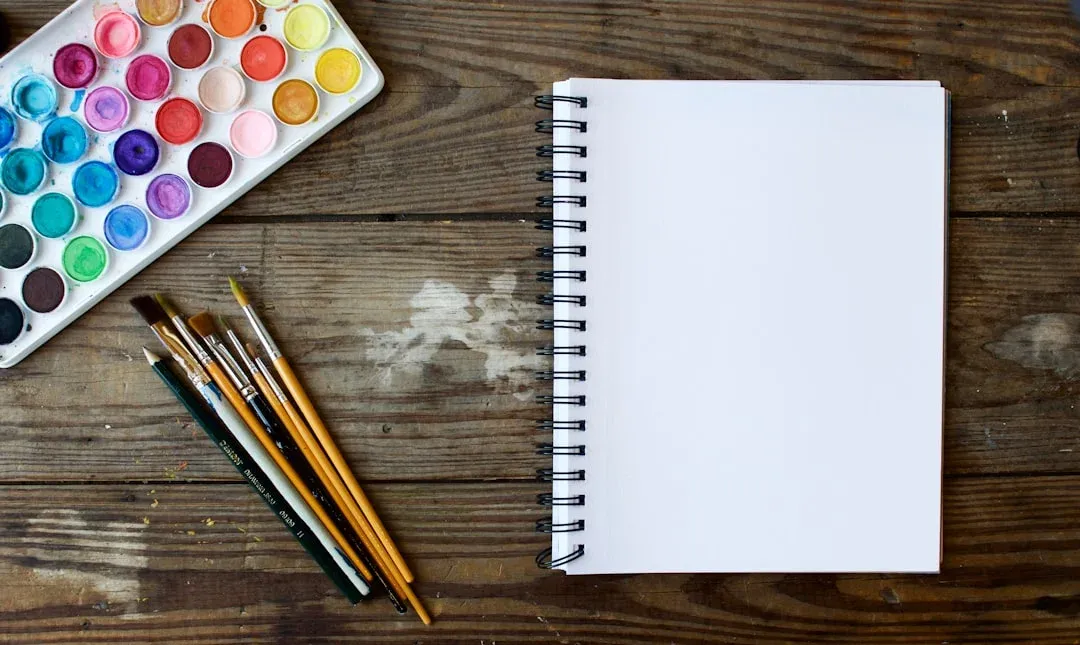 Watercolor paints, brushes, and a blank spiral notebook on a wooden table, ready for artistic use.