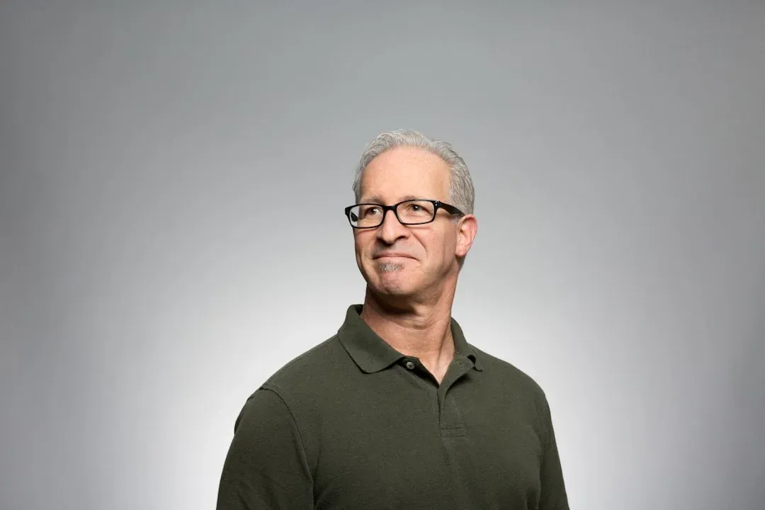 A man with short gray hair and glasses wearing a dark green polo shirt looks to the right against a plain gray background.