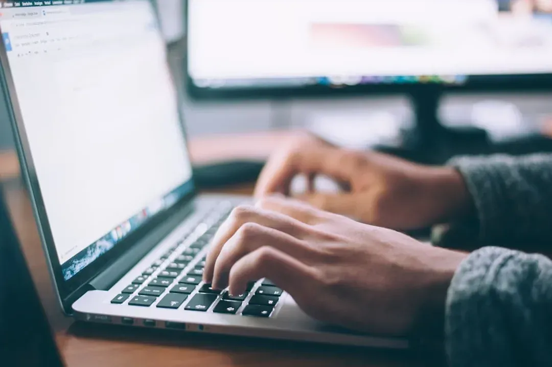 Person typing on a laptop with two monitors in the background, focused on a document on the screen.